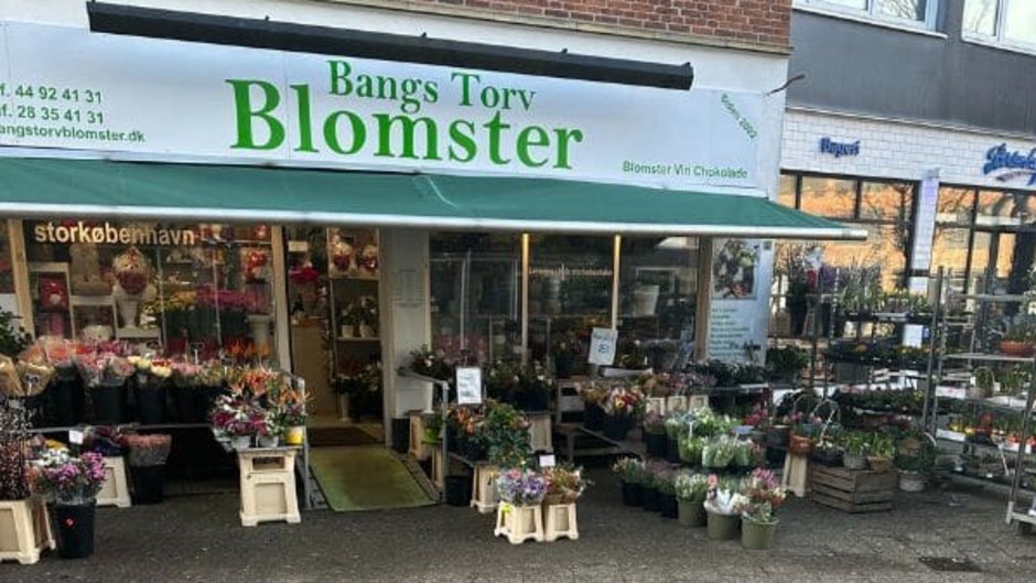 Billedet viser blomsterbutikken "Bangs Torv Blomster" med en facade i grøn og hvid. Udenfor er der flere blomsterbede og potter med blomster i forskellige farver, som pryder indgangen til butikken. Skyder og dekorative planter kan også ses langs facaden.